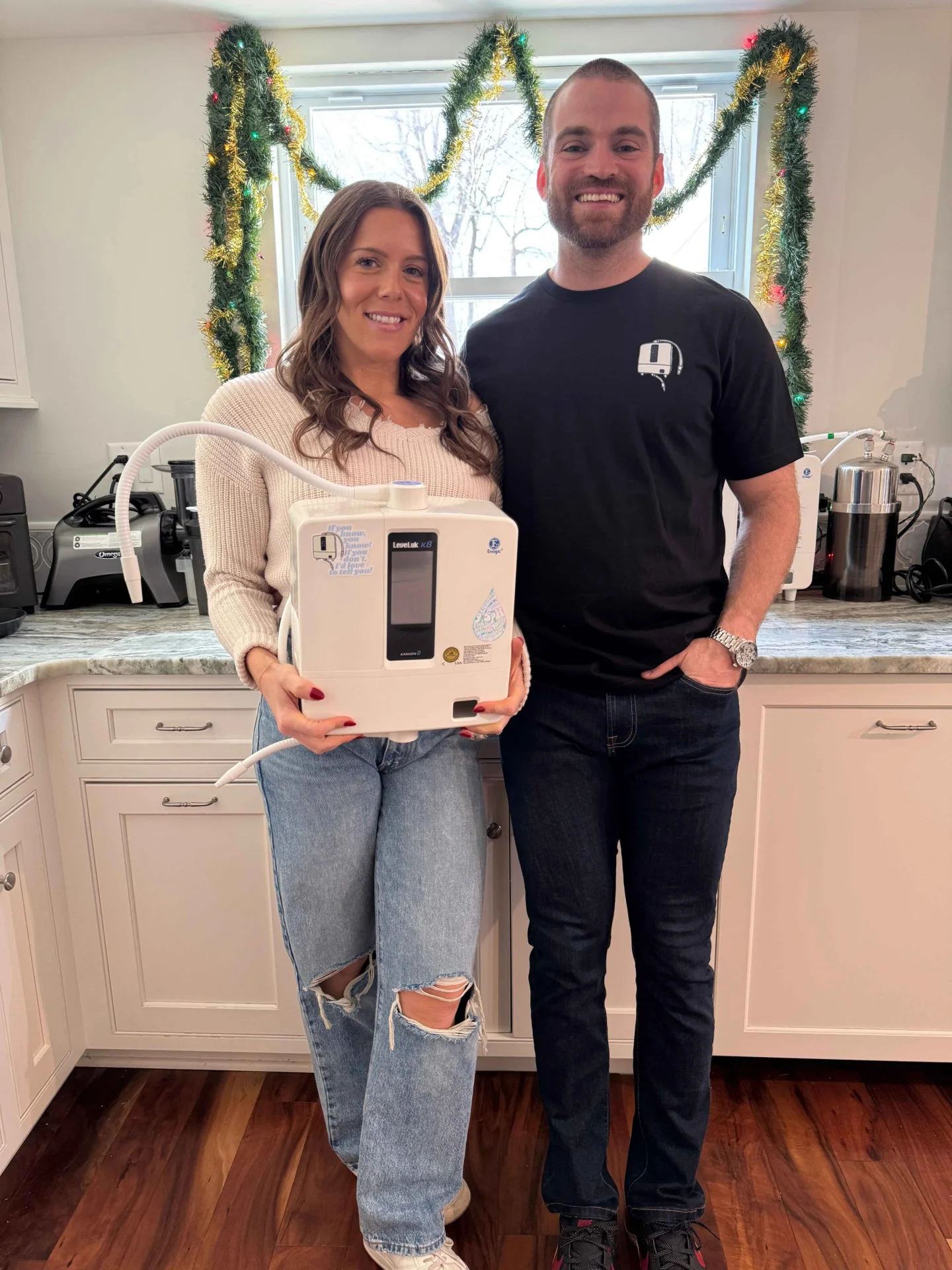 The Kangen Couple standing in a kitchen holding a new appliance box, with holiday garland in the background.