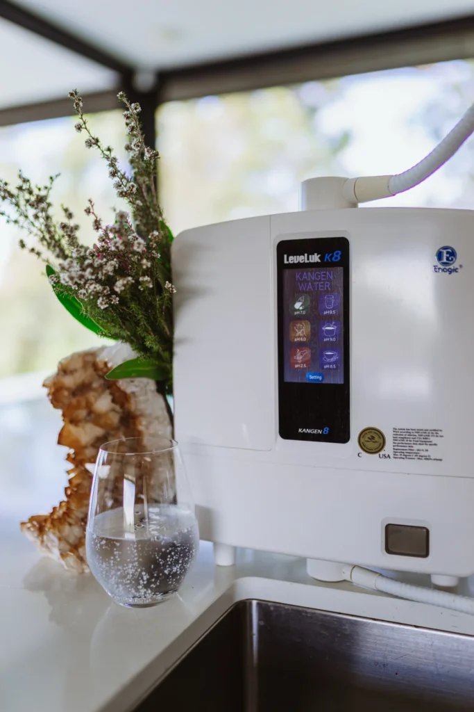 White Kangen water machine on a countertop next to a glass of water and a floral arrangement.