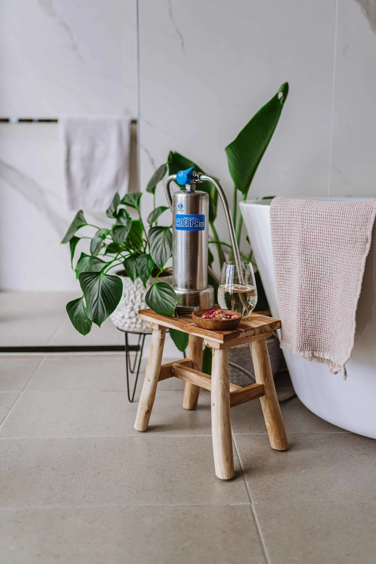 A stainless steel water filter is on a wooden stool beside a bath tub. A glass of water and a small bowl with snacks are next to the filter. There's a potted plant in the background.