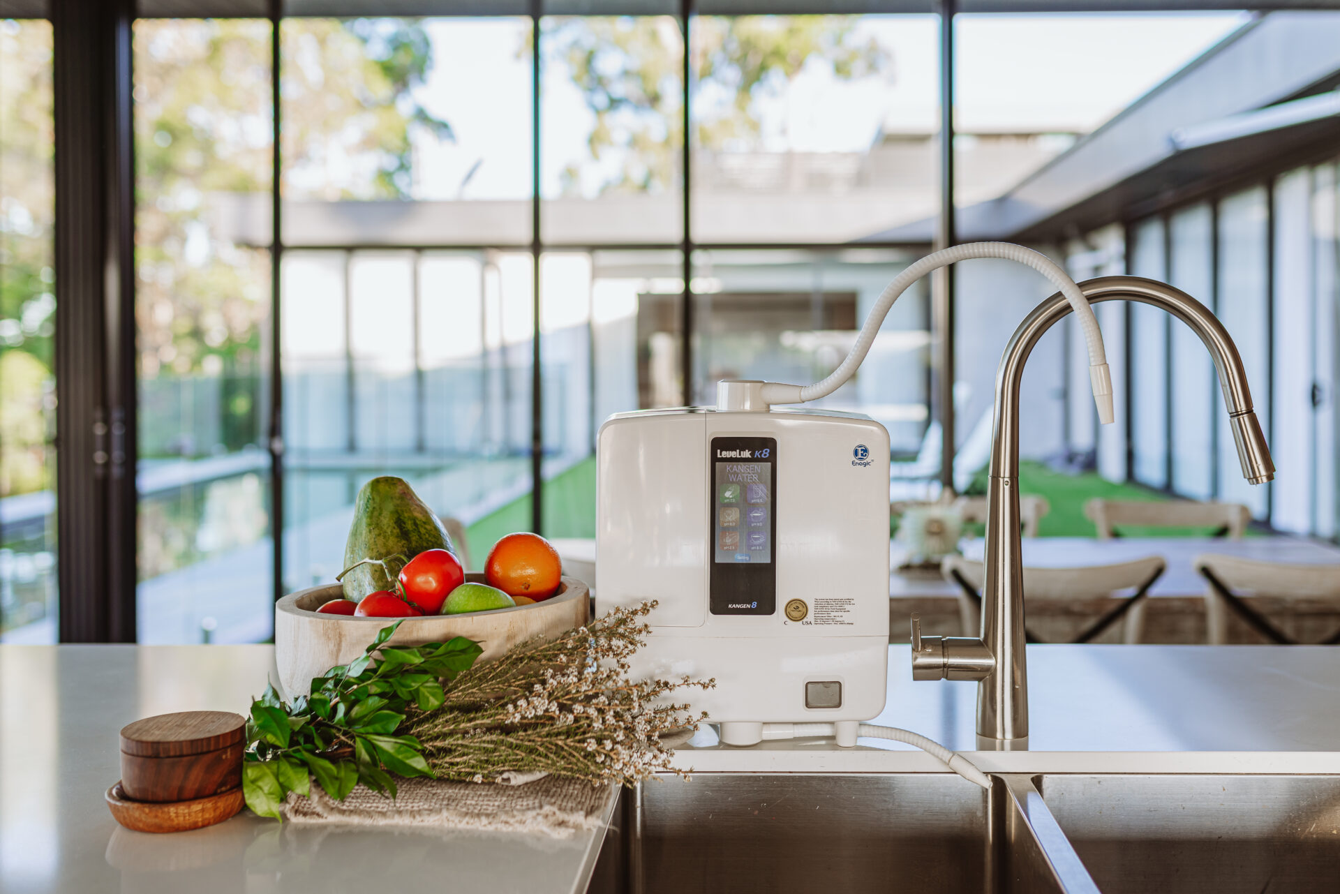 A water ionizer is placed on a modern kitchen counter near a sink, with a basket of fruits and herbs beside it, and a glass wall in the background showing a garden view.